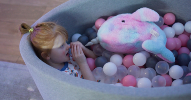 Minna sitting in a ball pit next to her narwal plush pillow, holding her hands up to her face.